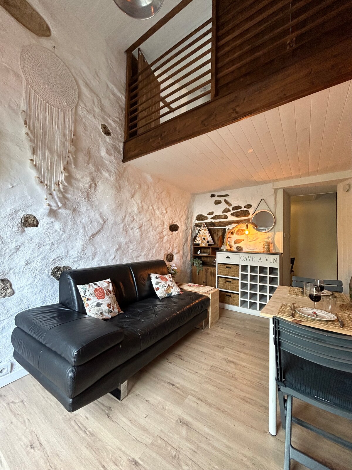 A living area is highlighted by a black sofa adorned with decorative pillows. The stone walls add texture, while a wooden wine rack and dining table enhance the space. Above, a loft area can be seen, adding a sense of openness and charm.