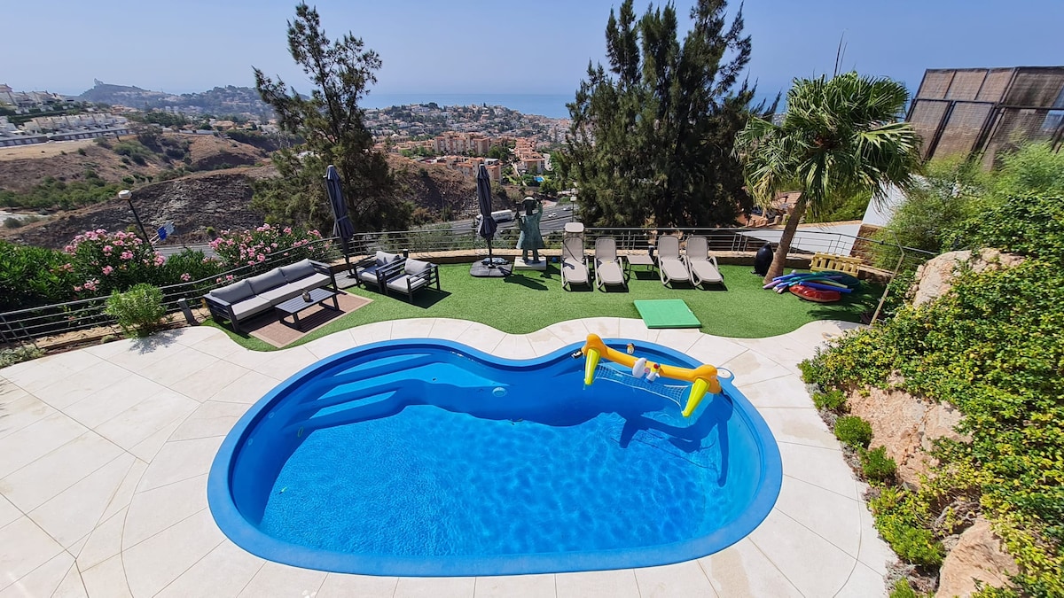 An inviting swimming pool is set in a spacious outdoor area, surrounded by loungers and shaded seating. Lush greenery and palm trees frame the scene, while distant views of the coastline provide a scenic backdrop. The area is well-suited for relaxation and enjoyment.