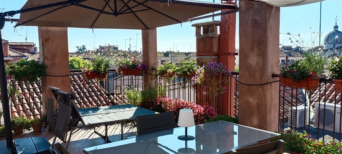 An outdoor terrace is displayed, featuring a dining table with an umbrella and six potted plants adorned with colorful flowers. The view includes rooftops of Venice and highlights a serene atmosphere, ideal for relaxation.