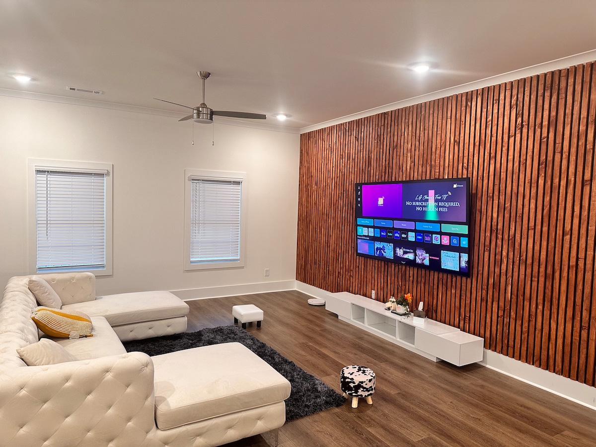 The living area features a modern sectional sofa positioned against a textured wooden accent wall. A large flat-screen TV is mounted, displaying a colorful interface. Natural light enters through two windows, complemented by subtle ceiling lighting and a plush area rug beneath the seating.