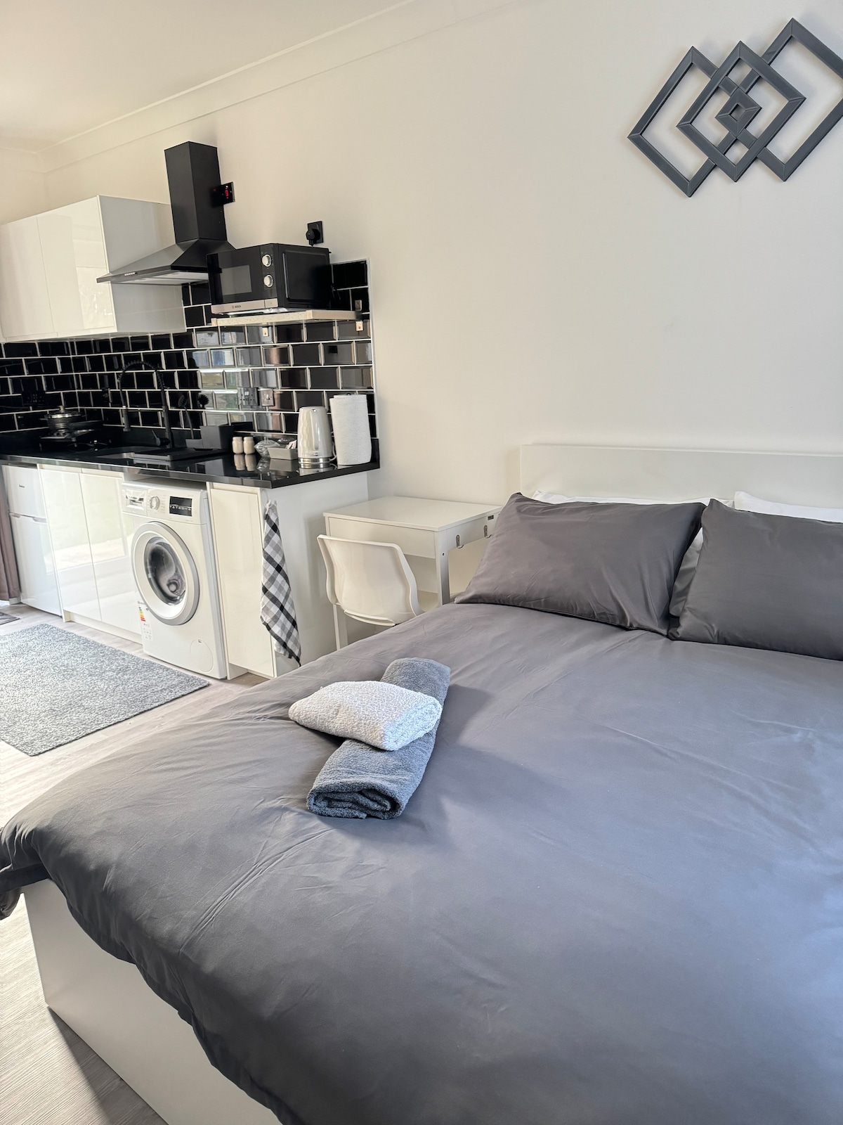 A studio space features a spacious bed covered with a grey duvet and towels neatly arranged on top. A small dining area with a chair is visible, along with a kitchenette equipped with modern appliances, including a washing machine and black tile backsplash.