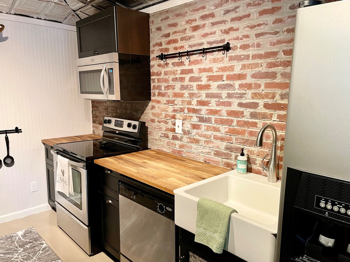 The kitchen features a combination of modern appliances and rustic charm, highlighted by a brick accent wall. Stainless steel appliances include an oven, microwave, and dishwasher, complemented by warm wooden countertops and a farmhouse-style sink.