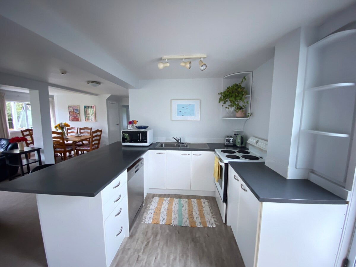 A modern kitchen is equipped with a range of appliances, including a microwave, stove, and refrigerator. Light cabinetry and a dark countertop create a cohesive look, complemented by a colorful area rug. The space flows into a dining area, visible in the background.
