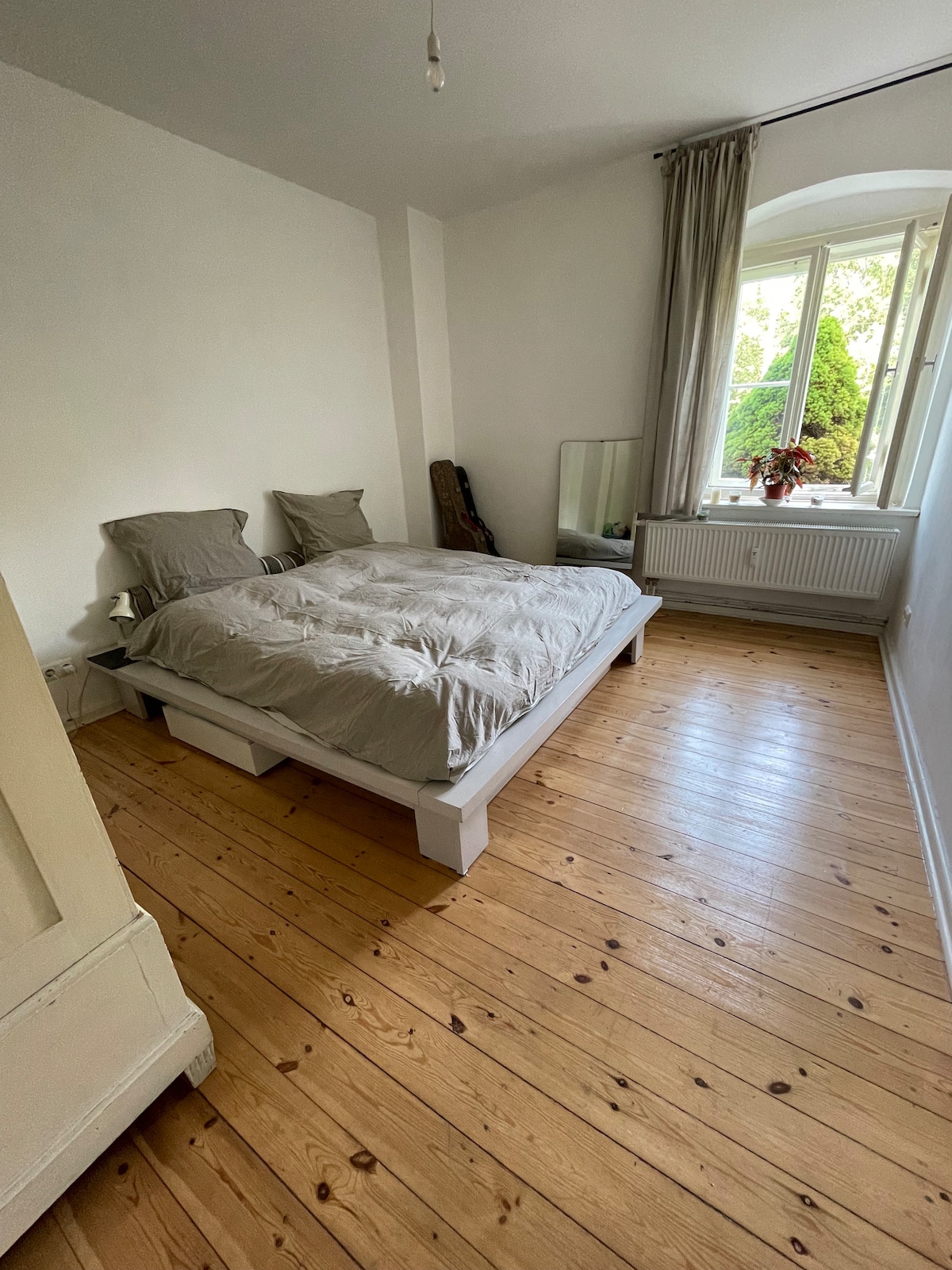The bedroom features a spacious bed dressed in light grey bedding, flanked by two pillows. Natural light enters through a large window, which is partially covered by soft curtains. A wooden floor adds warmth, complemented by a mirror and a decorative plant nearby.