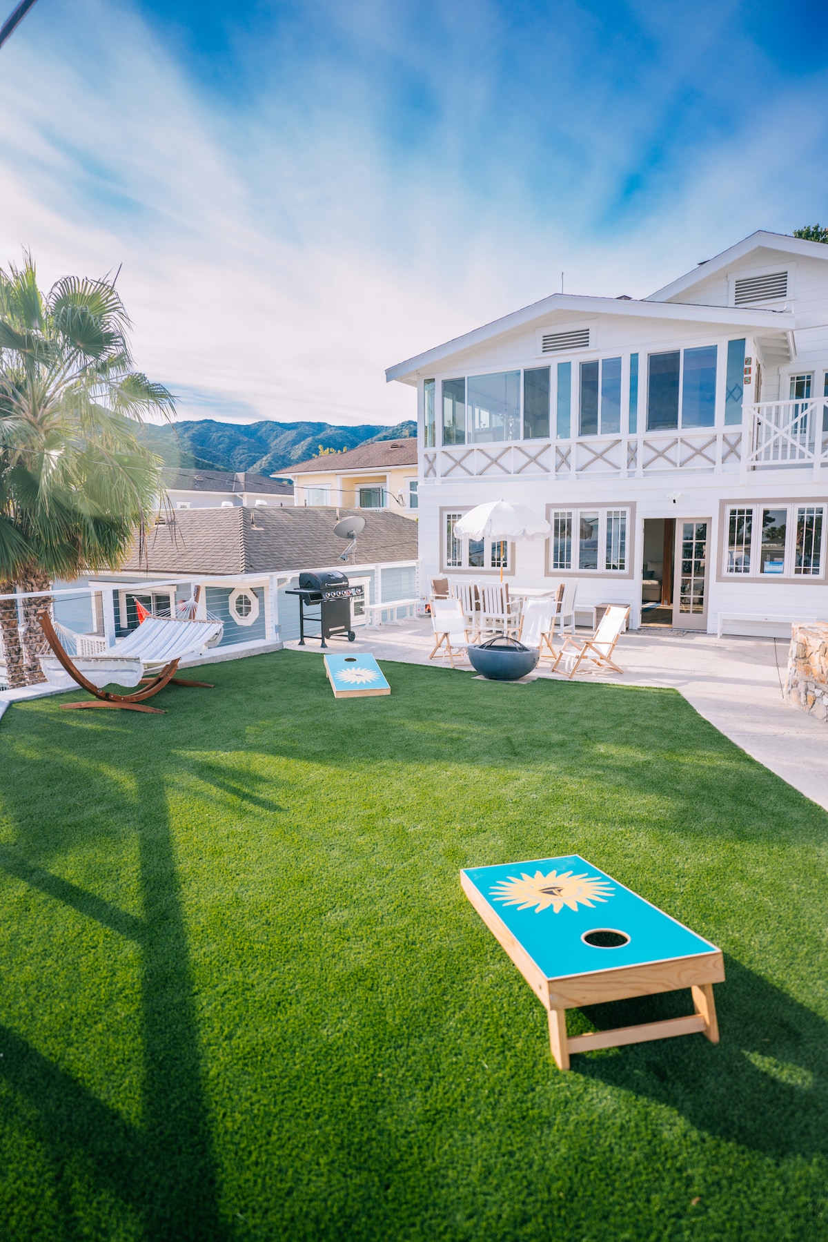 The outdoor yard features a lush, green lawn with cornhole boards set up for games. A hammock is visible nearby, along with a grill and several outdoor chairs, all set against a backdrop of the charming cottage and scenic mountain views.