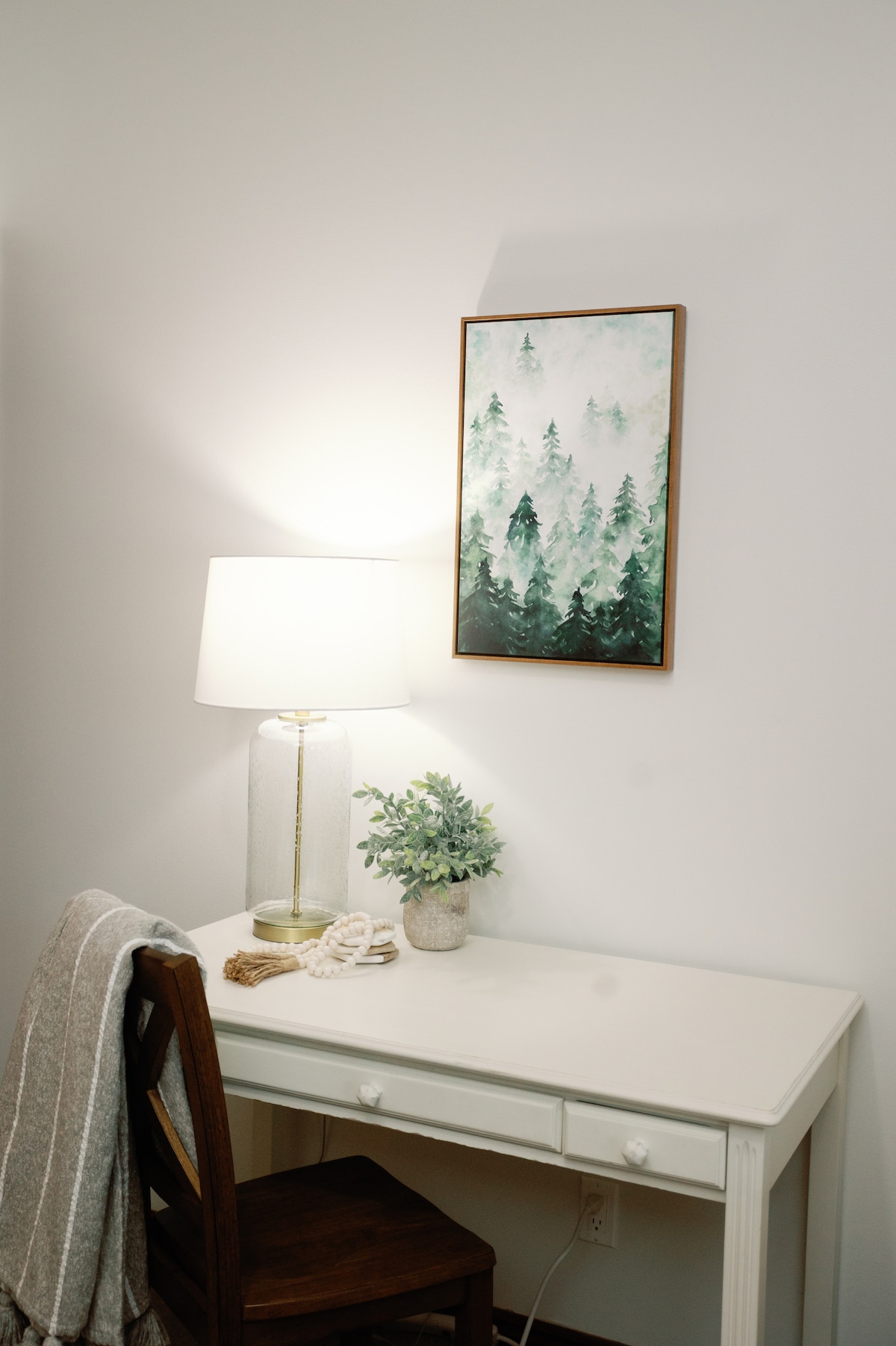 A small, white desk is positioned against a plain wall, accompanied by a wooden chair. A white lamp with a glass base illuminates the space. A framed artwork depicting a misty forest hangs above the desk, and a potted plant adds a touch of greenery.