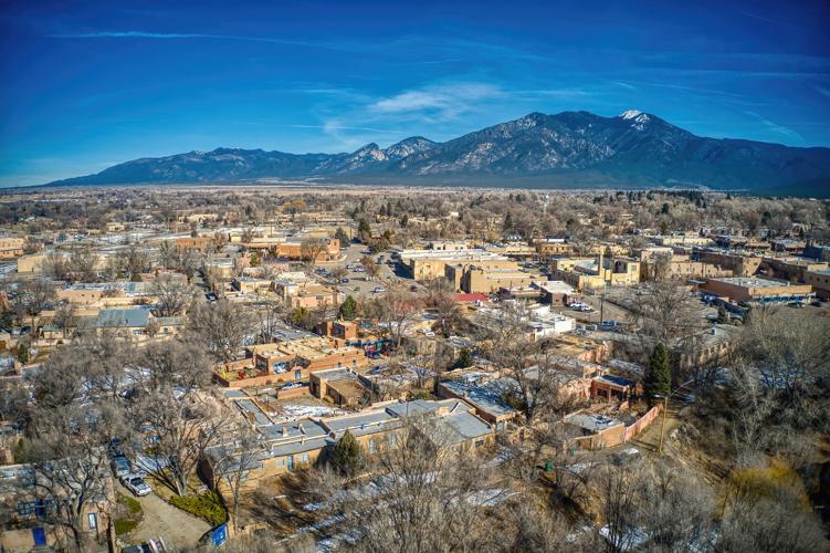 Aerial view of a town surrounded by mountains, featuring a mix of residential and commercial buildings. The landscape is characterized by bare trees and patches of snow, with the mountain range visible in the background under a clear blue sky.