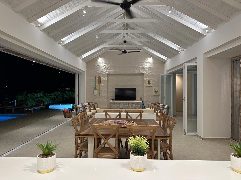 An open-concept living space features a large dining table surrounded by wooden chairs. A modern entertainment unit is situated on a white wall. Ceiling fans and recessed lighting provide a comfortable ambiance. Glass doors lead to an outdoor area with pool views, enhancing the sense of space.
