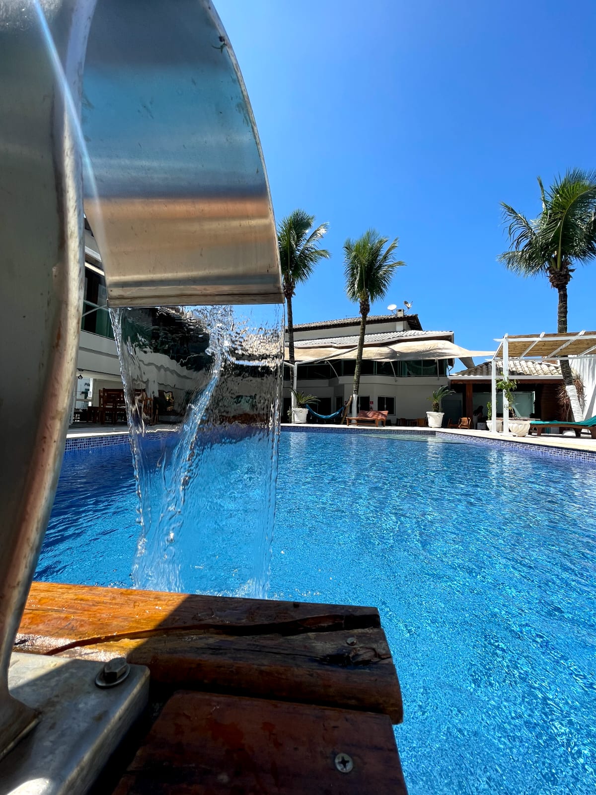 The image captures a close-up view of a waterfall feature pouring into a sparkling pool. Palm trees can be seen in the background against a clear blue sky, with lounge chairs positioned alongside the pool's edge. Sunlight reflects off the water's surface.