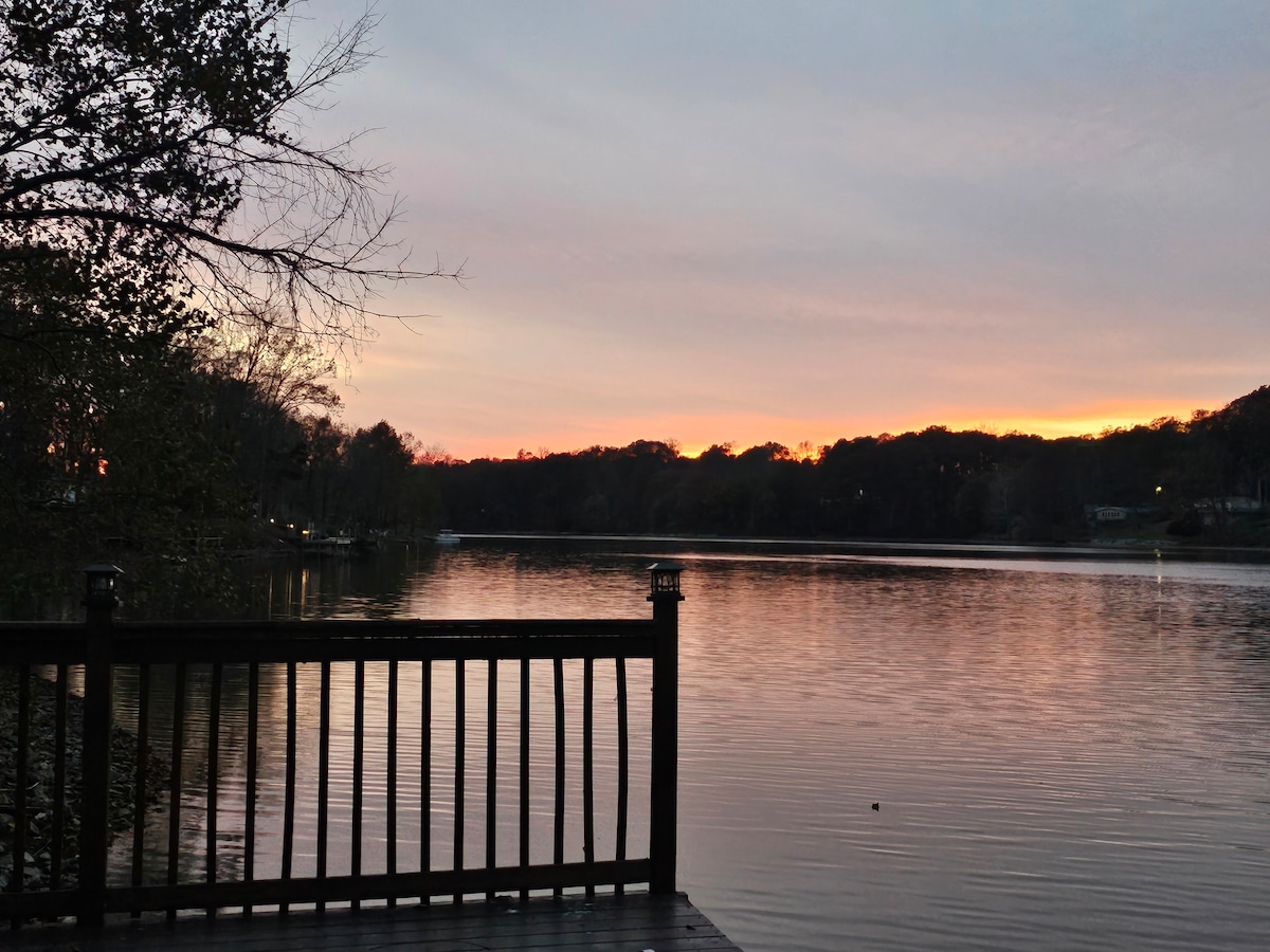 A calm lake is featured at dusk, reflecting soft hues of orange and pink from the sunset sky. A wooden deck with a railing overlooks the water, surrounded by trees and distant hills, creating a tranquil setting.
