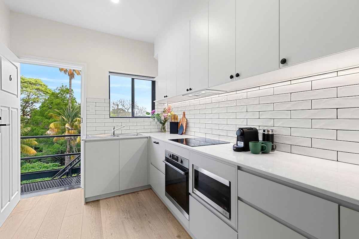 A modern kitchen features sleek cabinetry and white subway tile backsplash. Equipped with a refrigerator, oven, cooktop, and microwave, it offers ample counter space. A coffee pod machine and accessories are arranged neatly in view. Large windows provide natural light and scenic views of surrounding greenery.