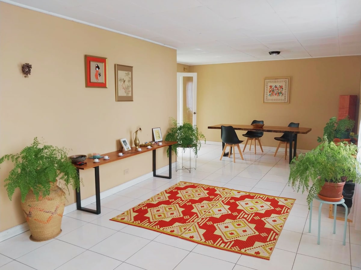 A spacious living area features a decorative area rug in warm tones, accented by green plants in various pots. A long table with black chairs is positioned in the background, while wall art adds subtle character to the beige walls.