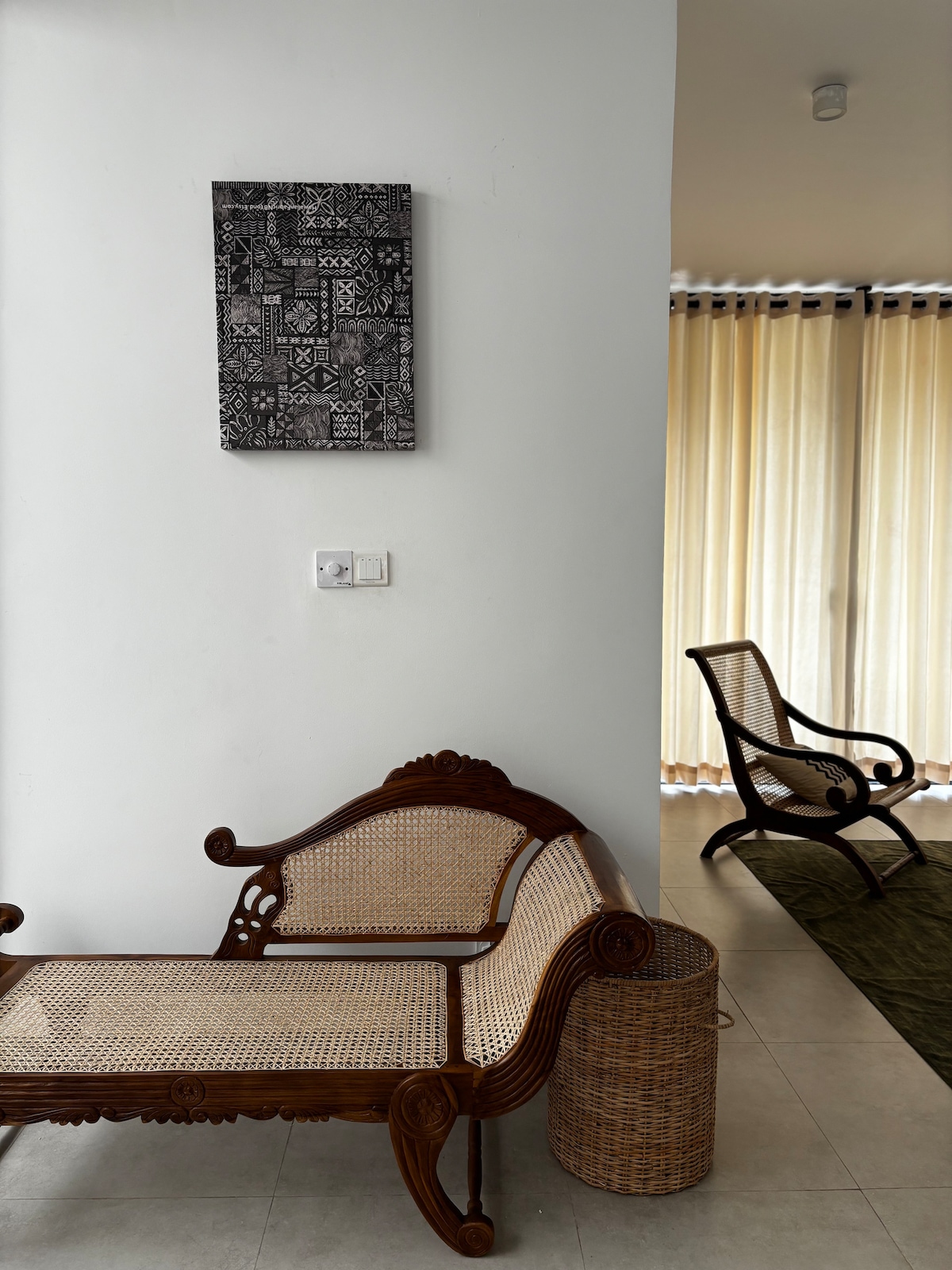 A serene seating area features intricately designed wooden furniture, including a curved bench and a woven chair. Natural light filters through sheer curtains, illuminating a decorative wall hanging with geometric patterns. A circular basket adds a functional touch to the space.
