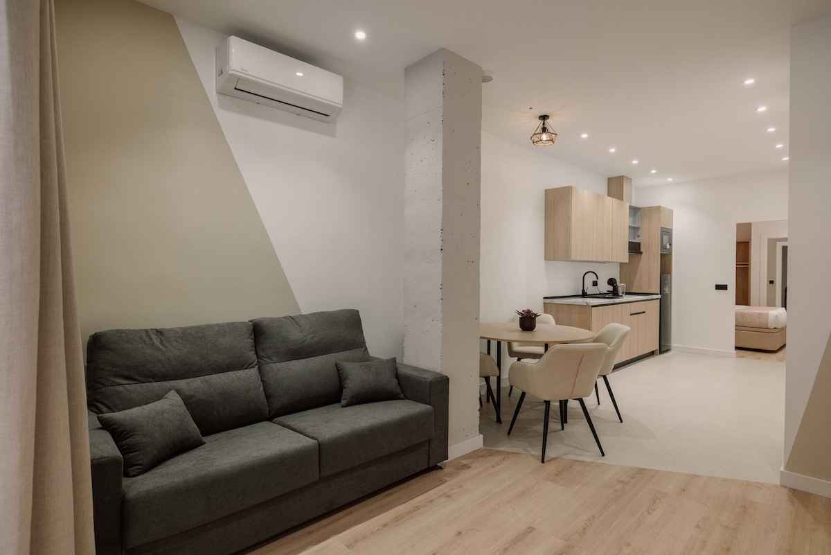 A modern living area features a spacious gray couch, complemented by a dining table and four chairs. Light wood flooring and soft lighting enhance the open layout, while a well-equipped kitchen with neutral cabinetry is visible in the background.