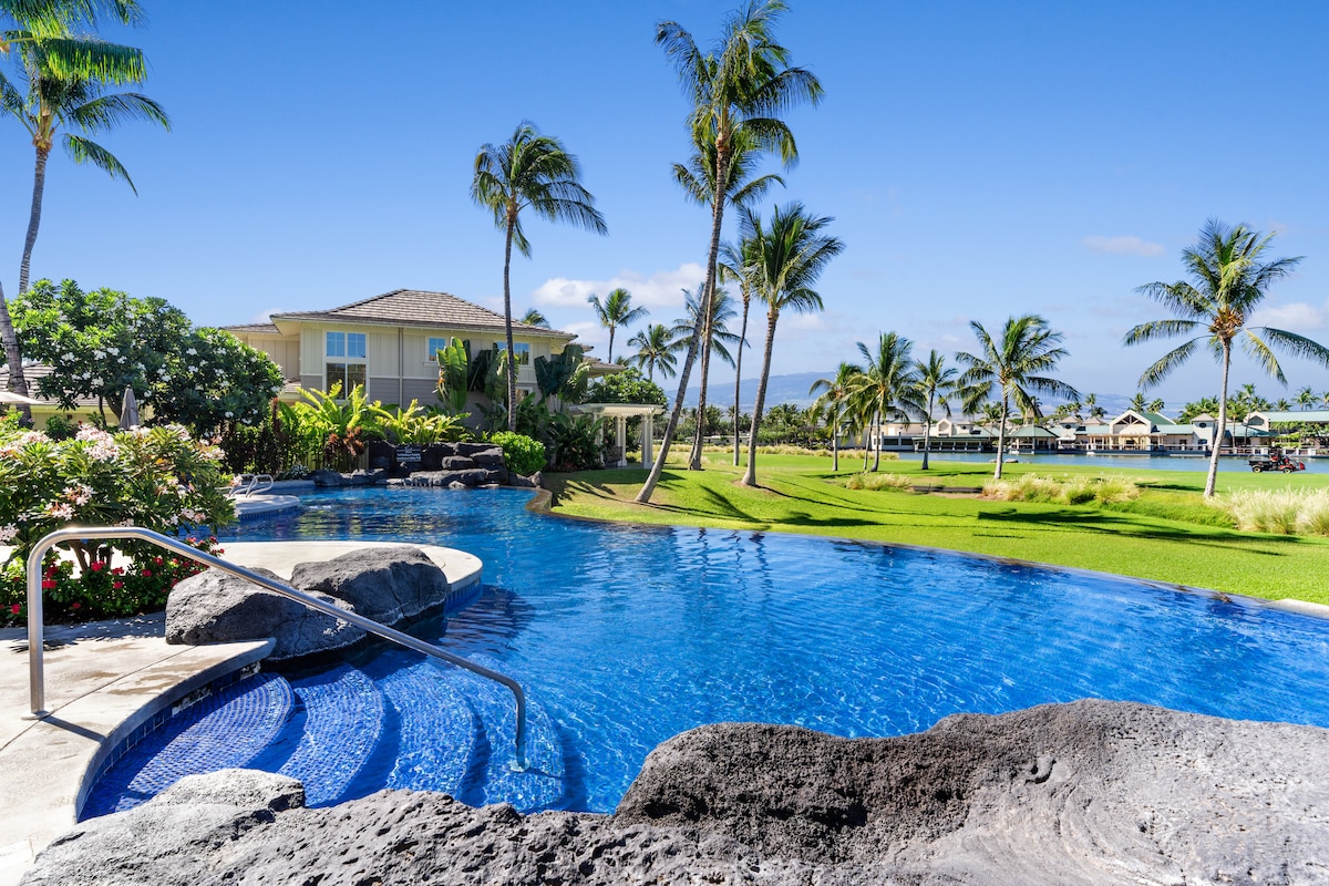 An infinity pool is framed by lush greenery and palm trees, providing a serene space for relaxation. The pool's gentle curves lead to stepped entry points, while a backdrop of well-maintained landscaping adds to the scenic view.