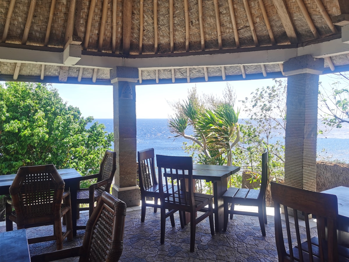 An open-air dining area features wooden tables and chairs, framed by tropical greenery. The ocean can be seen in the background, with gentle waves visible on the horizon, enhancing the serene atmosphere. The thatched roof provides shade while allowing for a refreshing breeze.