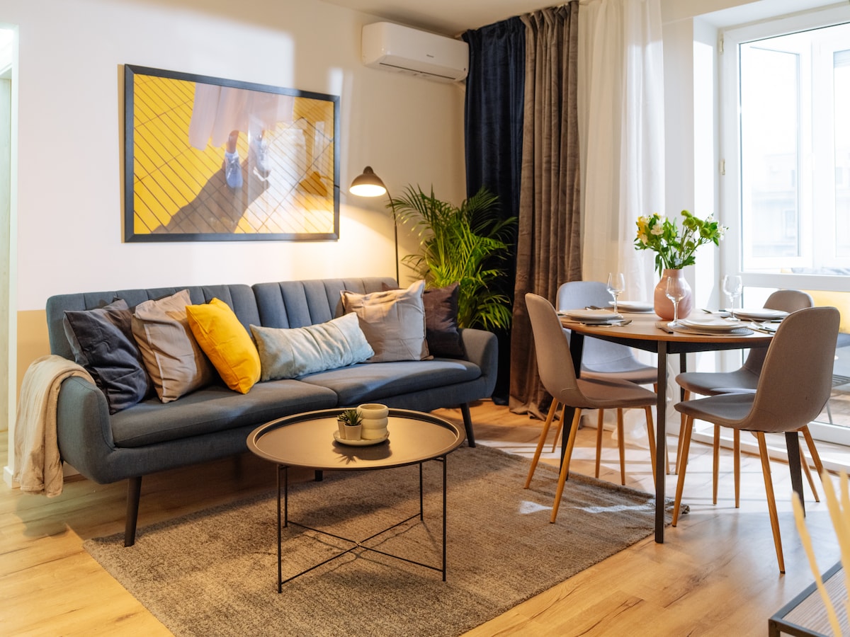 A modern living area features a stylish blue sofa adorned with various decorative pillows. A round table set for dining is visible with elegant dishware, while a floor lamp provides soft illumination. A potted plant adds a touch of greenery, and a window allows natural light to enter.