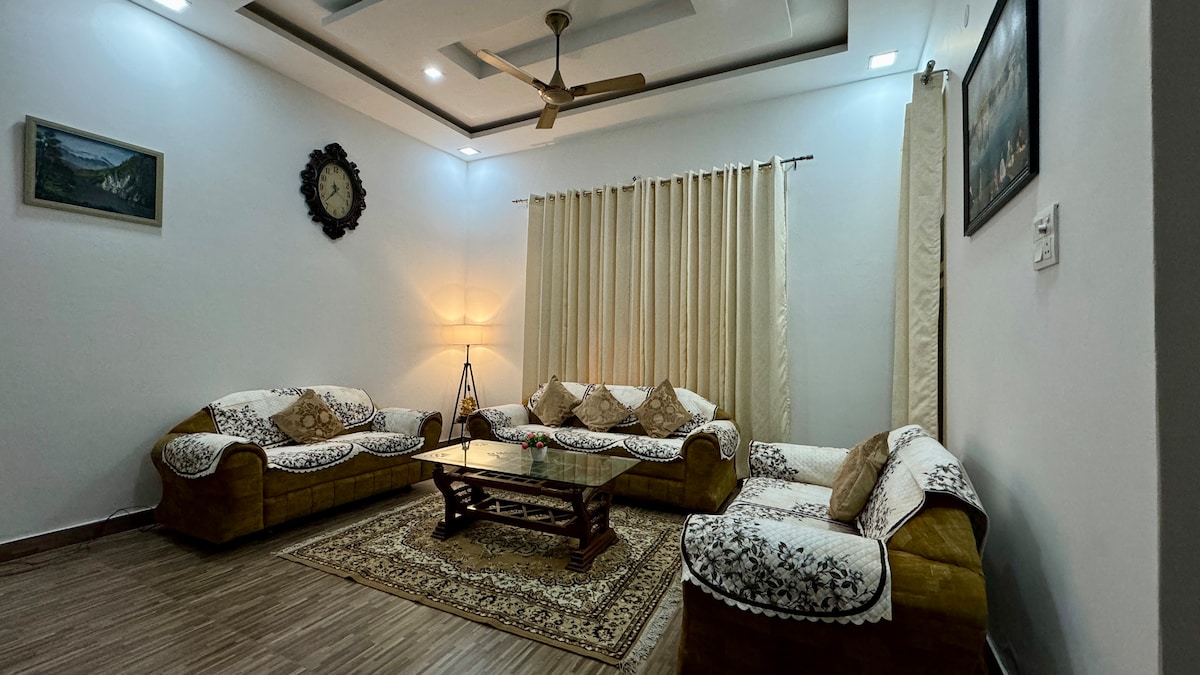 A cozy living room features a large area rug centered under a wooden coffee table. Two comfortable sofas are adorned with decorative cushions. Soft lighting is provided by a floor lamp, while curtains frame the windows. A clock and framed artwork add subtle touches to the decor.