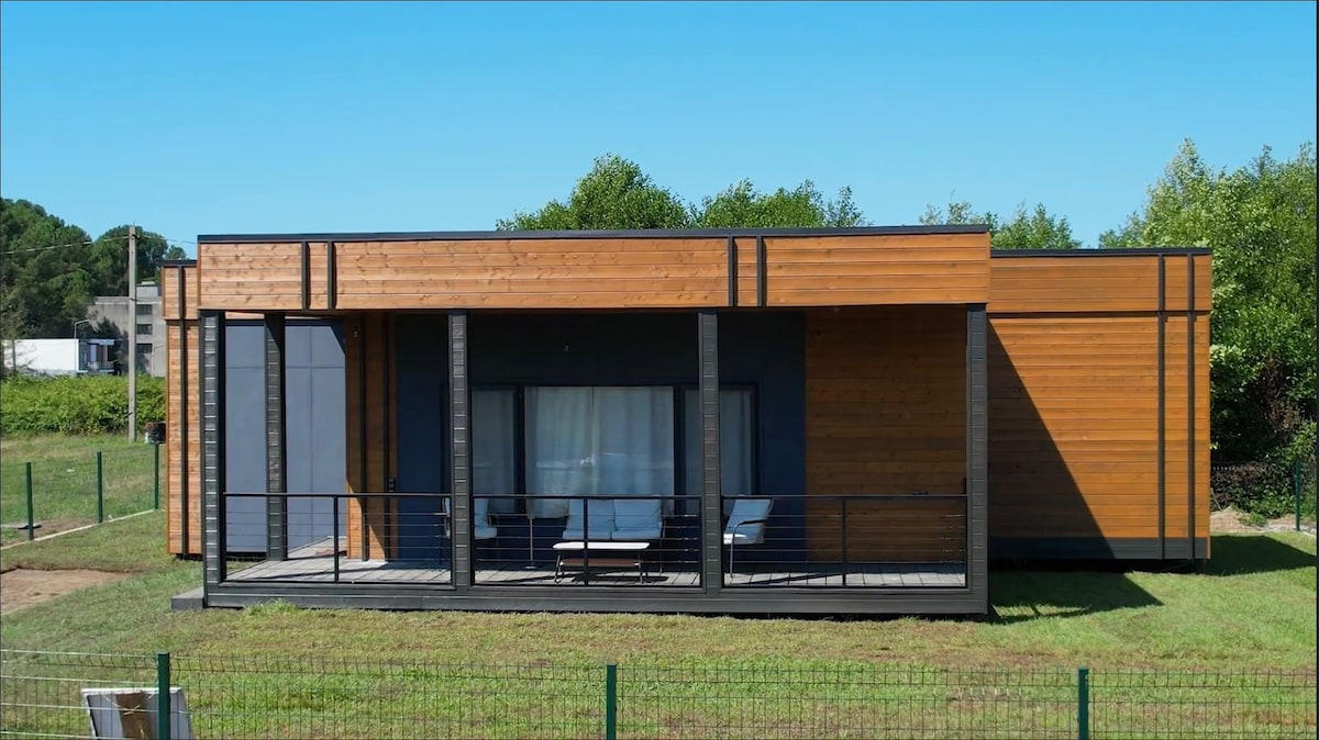 A modern wooden structure features a spacious porch with seating, providing a connection to the outdoors. Large windows allow natural light to fill the interior, while green grass and trees surround the home, enhancing the tranquil setting.