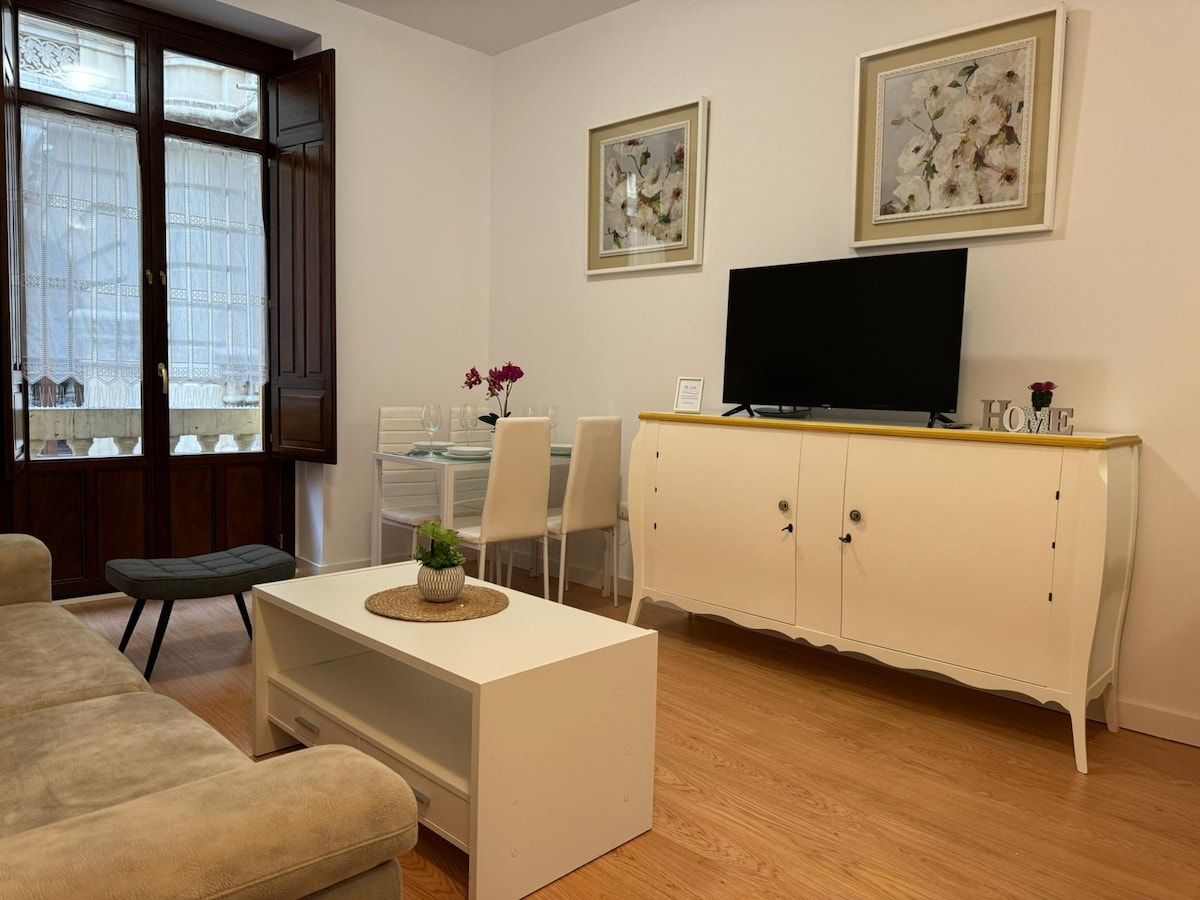 A living area features a light-colored sofa and a low coffee table. A modern television is positioned on a stylish white cabinet. In the background, a dining table with transparent chairs is set against large windows allowing natural light to brighten the space.