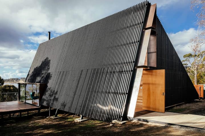  The image shows a sleek, modern A-frame cabin with a black slatted exterior and sharp angles. A small wooden deck extends from the side, complementing the minimalist wooden entrance. The bold design contrasts beautifully with the natural surroundings.
