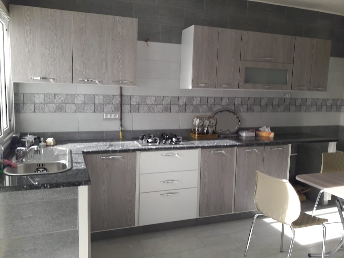 A modern kitchen is equipped with wooden cabinetry and a dark countertop, offering ample storage and workspace. A sink and stove are positioned on one side, while a dining table with several chairs is placed nearby. Natural light enters through a large window.