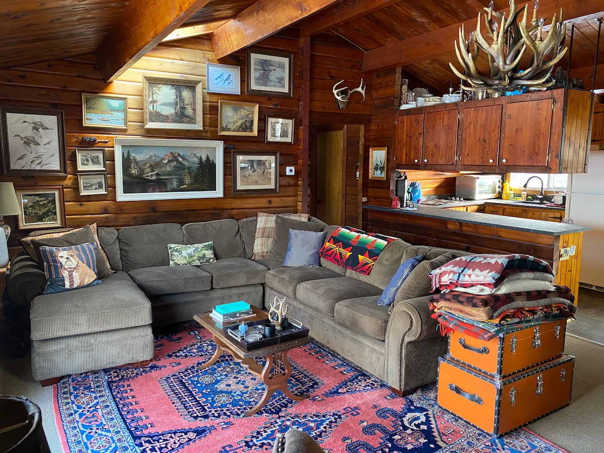 A cozy living area is depicted, featuring a large sectional sofa adorned with colorful pillows and throws. The wooden walls are decorated with framed artwork and photographs. A coffee table sits in front of the sofa, while an open kitchen area is visible in the background.