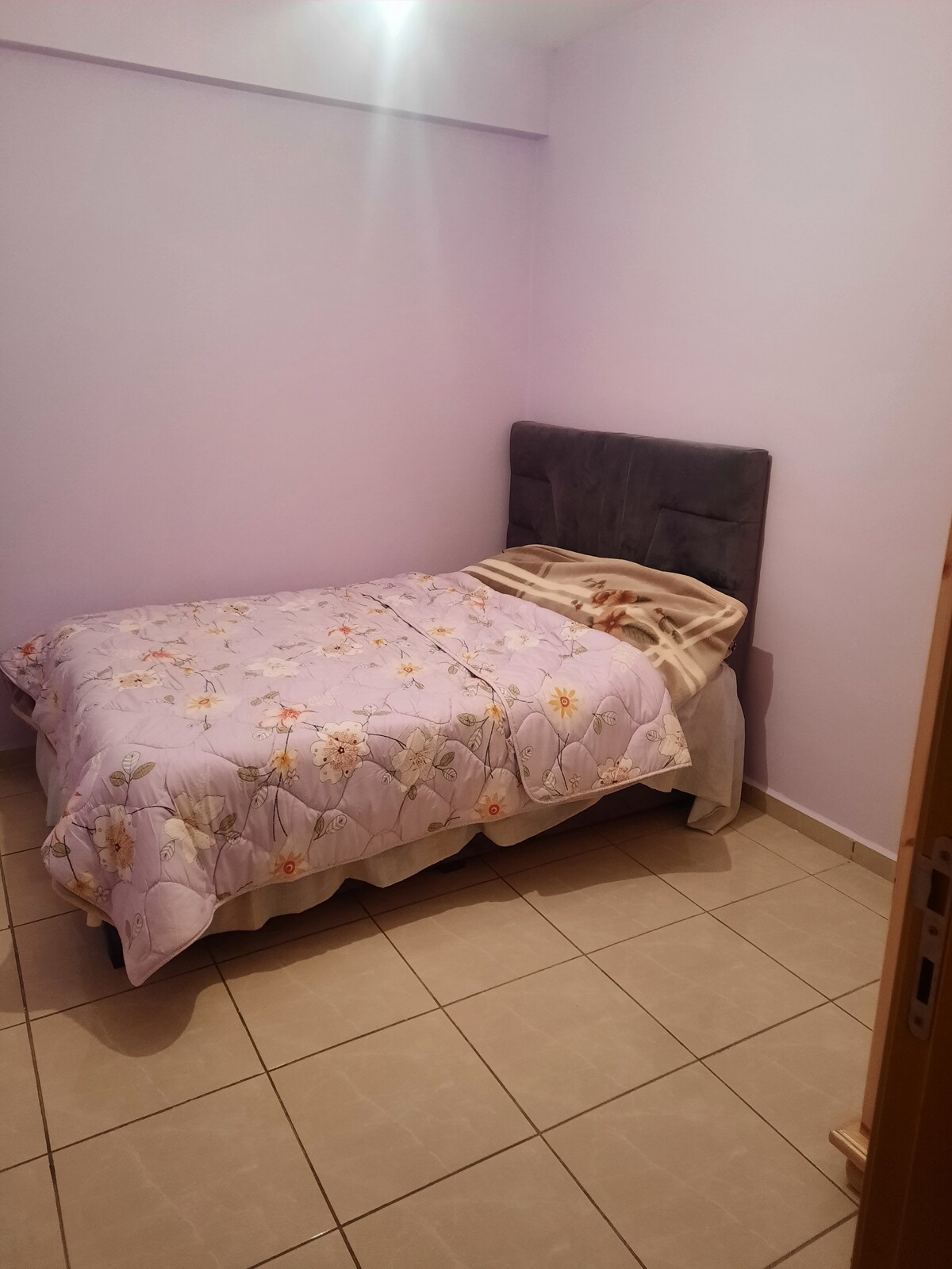 A simple sleeping area features a spacious bed covered with a floral patterned blanket. Light purple walls create a soft ambiance, while neutral-colored tiles contribute to the overall calmness of the room.