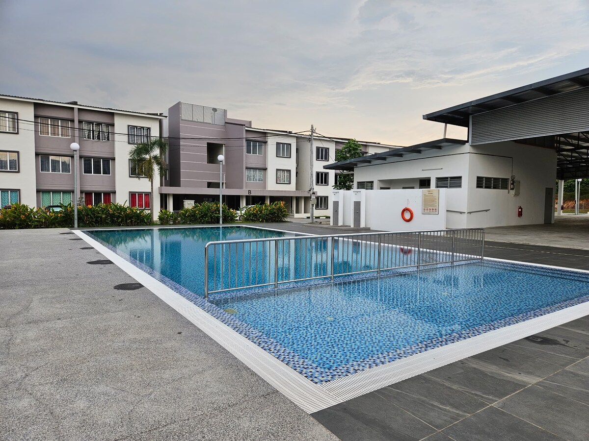 A spacious swimming pool is situated in a well-maintained outdoor area, surrounded by lush greenery. The pool features a rectangular shape with inviting turquoise water, and safety rails are installed along one side, ensuring accessibility. The surrounding architecture includes multi-story buildings for added context.