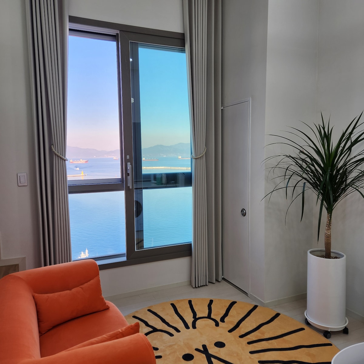 A corner of the living space features a large window that provides sweeping views of the tranquil sea and distant mountains. An orange couch sits nearby, complemented by a patterned area rug, while a tall plant adds a touch of greenery.
