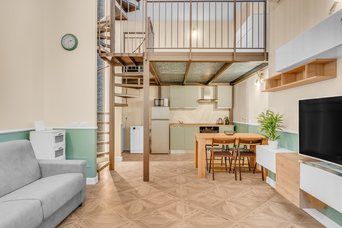 An open living area features a spiral staircase leading to a loft. A well-equipped kitchen is visible alongside a dining table with four chairs. A comfortable gray sofa is positioned nearby, and a flat-screen TV is mounted on the wall opposite the kitchen.
