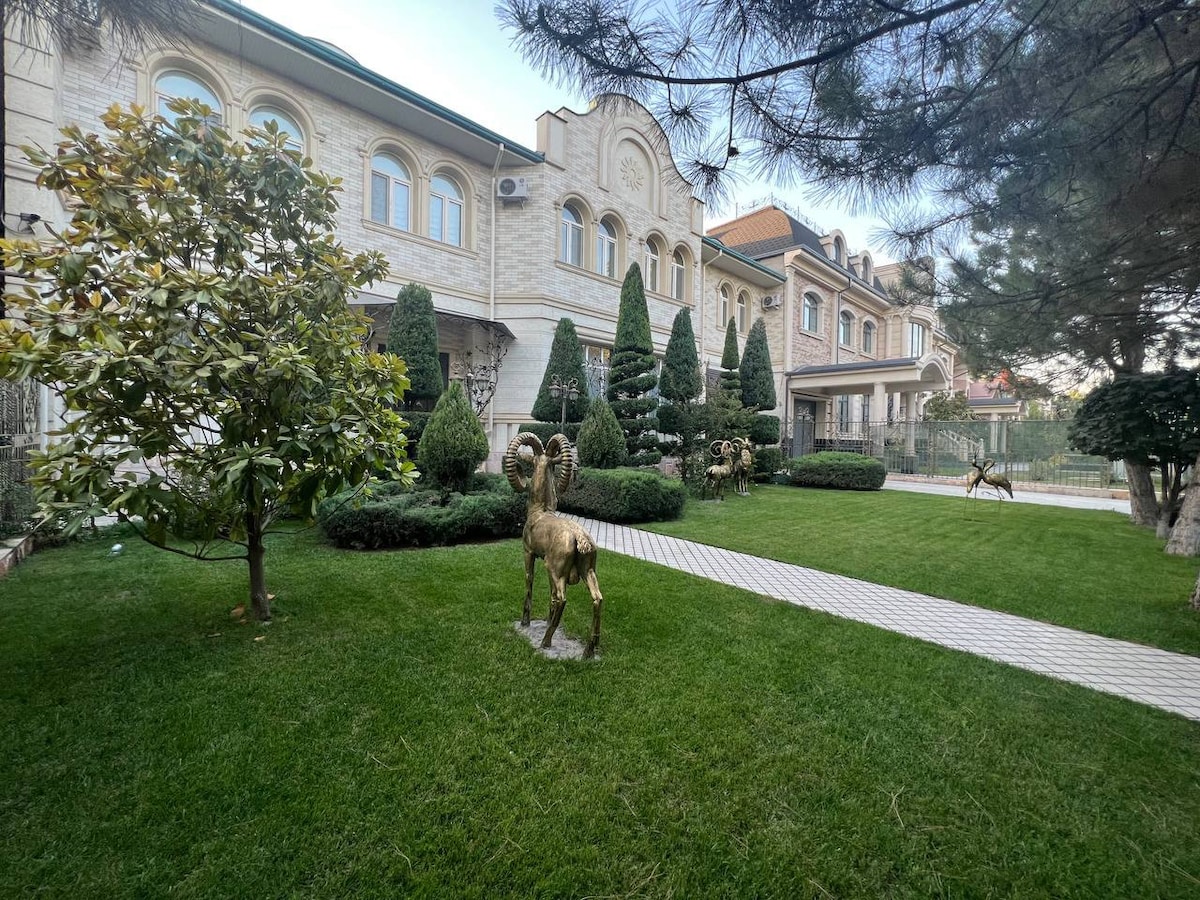 A manicured lawn features sculpted bushes and ornamental trees, creating a serene outdoor space. Bronze sculptures of rams are positioned gracefully across the yard, which is bordered by a charming pathway leading to a grand stone residence.