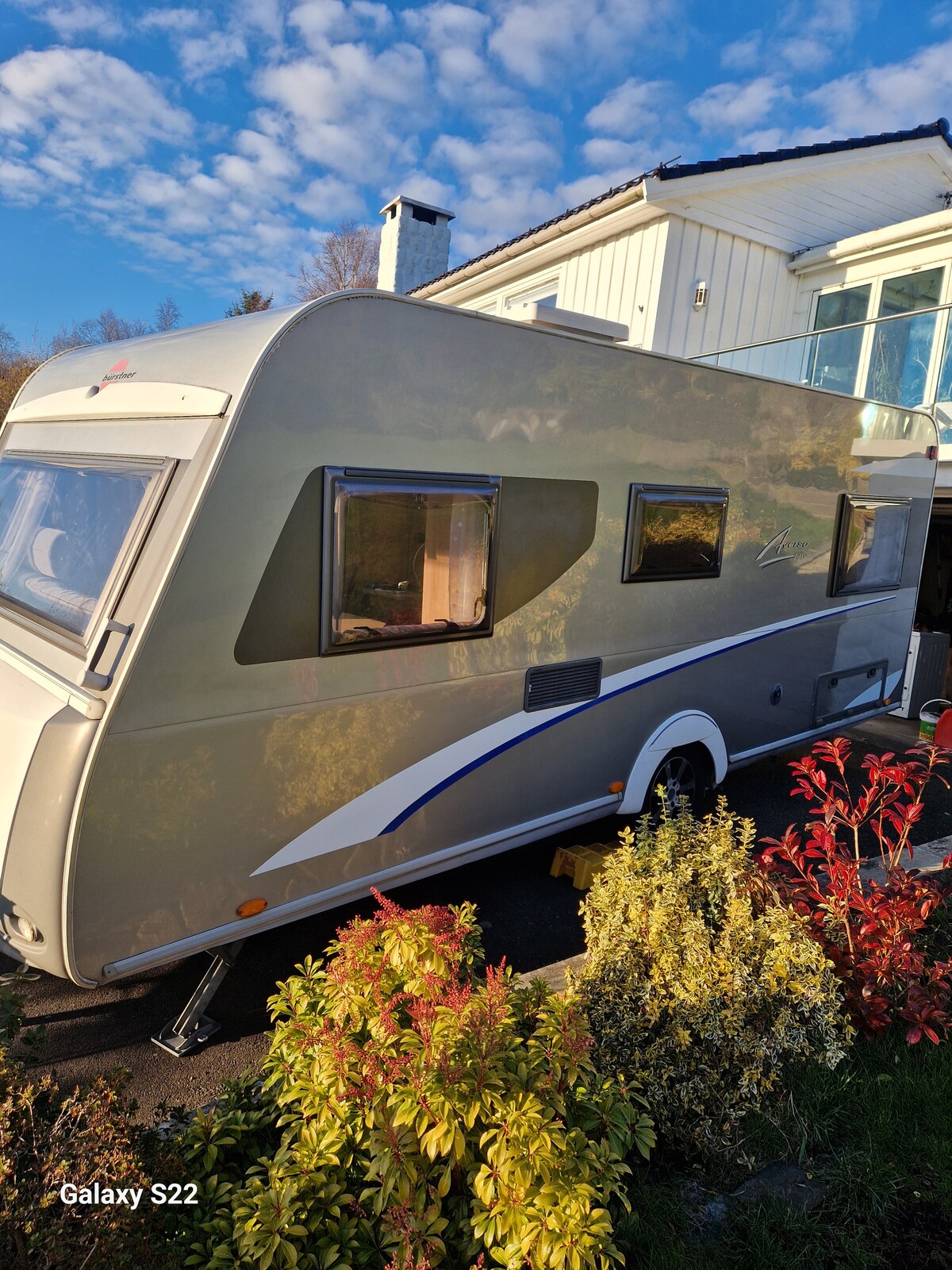 A sleek camper trailer is parked beside a light-colored house. The trailer features large windows and a contemporary design, with a white and gray exterior accented by subtle blue detailing. Lush greenery and colorful foliage are visible in the foreground.
