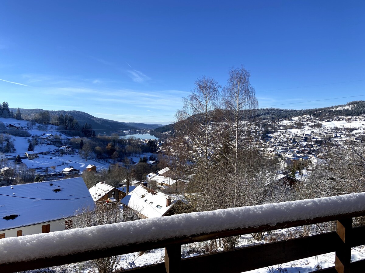 A winter landscape features a panoramic view of snow-covered rooftops nestled within hills. The lake glistens in the distance under a clear blue sky, while a wooden railing frames the scene, adding warmth to the cold setting.