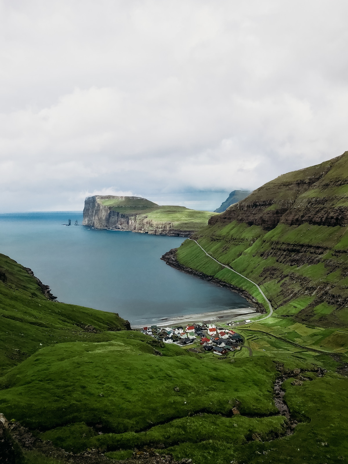 A panoramic view of Tjørnuvík is captured, showcasing a small village nestled between lush green hills and the Atlantic Ocean. Towering cliffs rise dramatically nearby, while the coastline curves gently, revealing the village's colorful rooftops against the vibrant green landscape and cloudy sky.