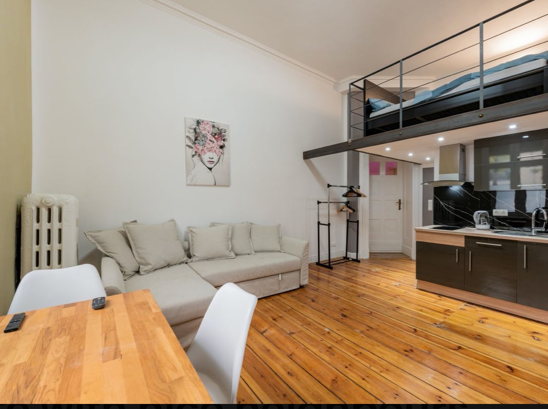 A spacious living area features a light-colored sofa and a wooden dining table. An open kitchenette is visible, equipped with modern appliances. Above, a gallery sleeping area is accessed by a sleek black railing. Natural light enters through the window, highlighting the wooden floor.