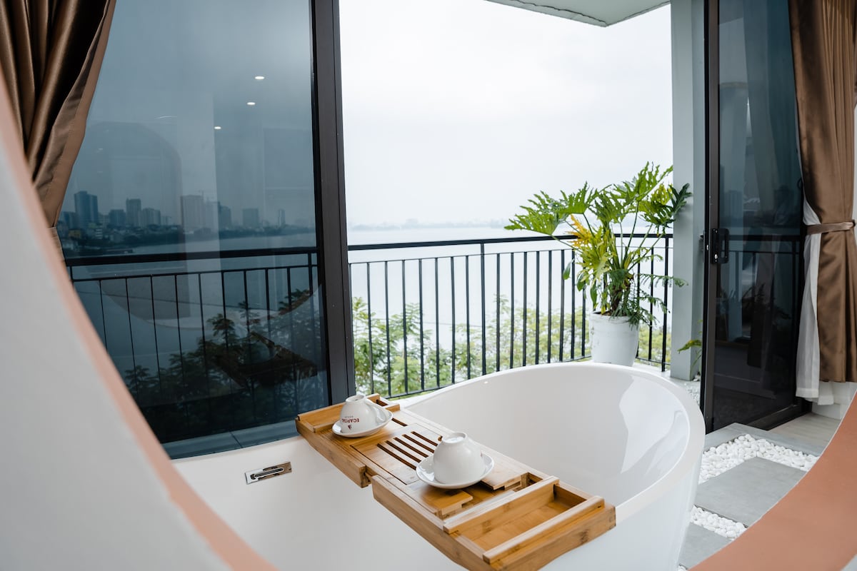 A modern freestanding bathtub is positioned near expansive glass doors, offering a view of the lake and city skyline. A wooden bath tray is placed across the tub, accommodating tea cups. Lush greenery is visible outside, with a balcony railing enhancing the scenic perspective.