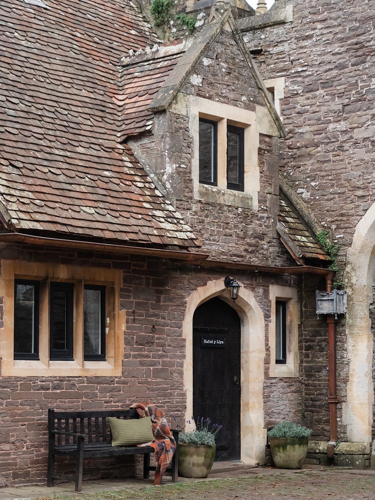 The cottage exterior features rustic stone walls and a sloping roof with wooden shingles. A dark wooden door is set within an archway, flanked by tall windows. A wooden bench adorned with cushions sits on the cobbled pathway, enhanced by potted lavender plants nearby.
