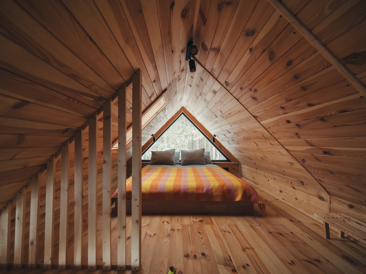An inviting loft space showcases a queen-size bed with a colorful blanket, framed by wooden walls and a sloped ceiling. Large triangular windows provide views of the surrounding forest, allowing natural light to brighten the room.
