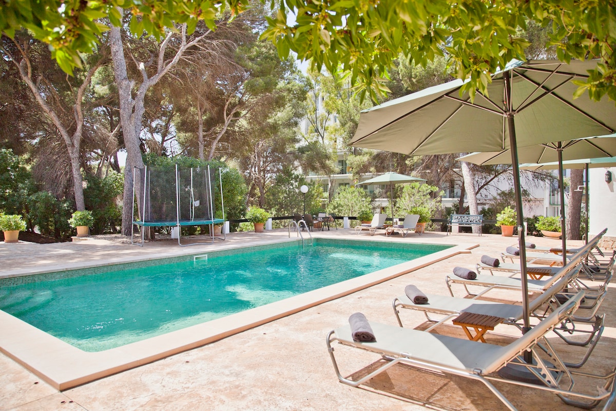 An outdoor area features a large pool surrounded by sun loungers with rolled towels. Umbrellas provide shade, and a trampoline is visible in the background. Lush greenery and plant pots enhance the serene environment. The space offers a relaxed setting for family enjoyment.