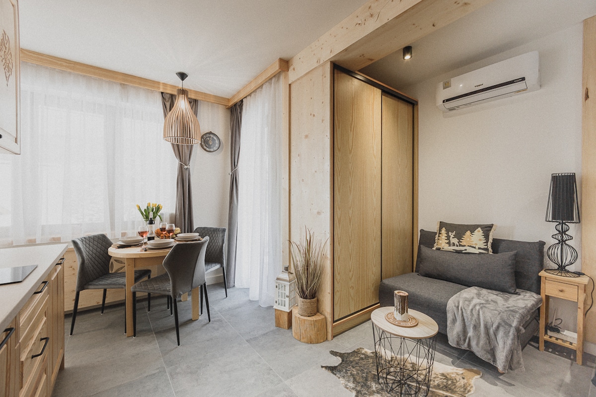 A cozy studio apartment features a dining area with a round table and chairs, a comfortable sofa, and wooden accents. A wall-mounted air conditioner is installed for climate control. Natural light filters through sheer curtains, highlighting the inviting interior design.