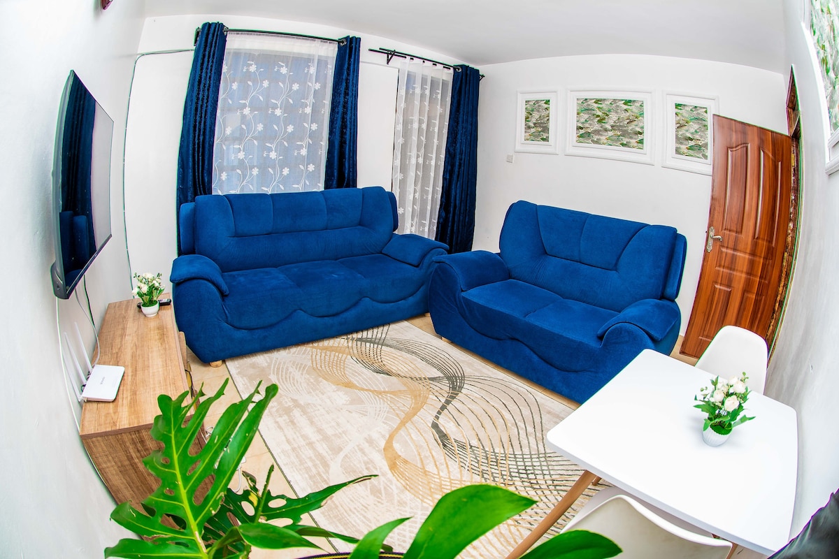 A bright living area features two spacious blue sofas facing a wall-mounted television. Natural light enters through sheer curtains, illuminating a light-colored rug with elegant patterns. A small white dining table sits nearby, accompanied by a decorative plant on the table.