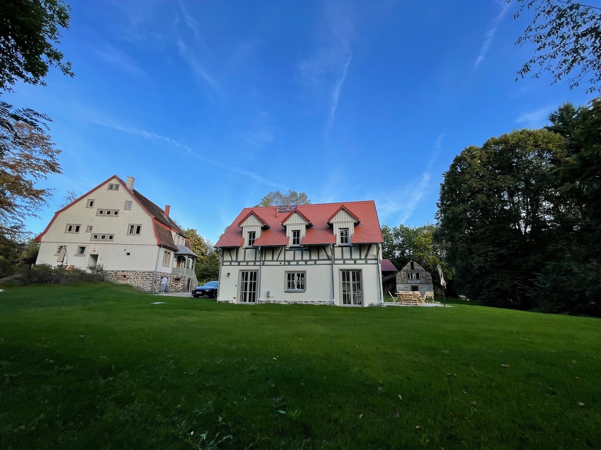 A charming structure is set against a clear blue sky, featuring a mix of traditional and modern architectural elements. The building is bordered by lush green lawns and surrounded by trees, providing a serene outdoor setting.