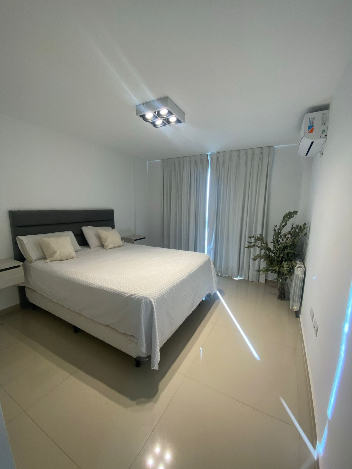 A well-lit bedroom features a spacious bed with two decorative pillows. Large windows are draped with light curtains, allowing natural light to filter in. A small plant adds a touch of greenery, while the polished floor reflects the ambient lighting.