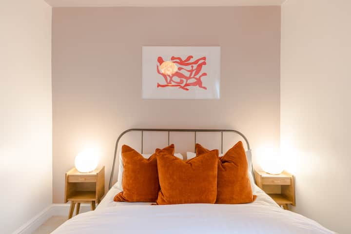 Opustite se u mirnoj spavaćoj sobi s udobnim bijelim krevetom, narančastim jastucima, mekom rasvjetom i umirujućim umjetničkim djelima za mir i udobnost.