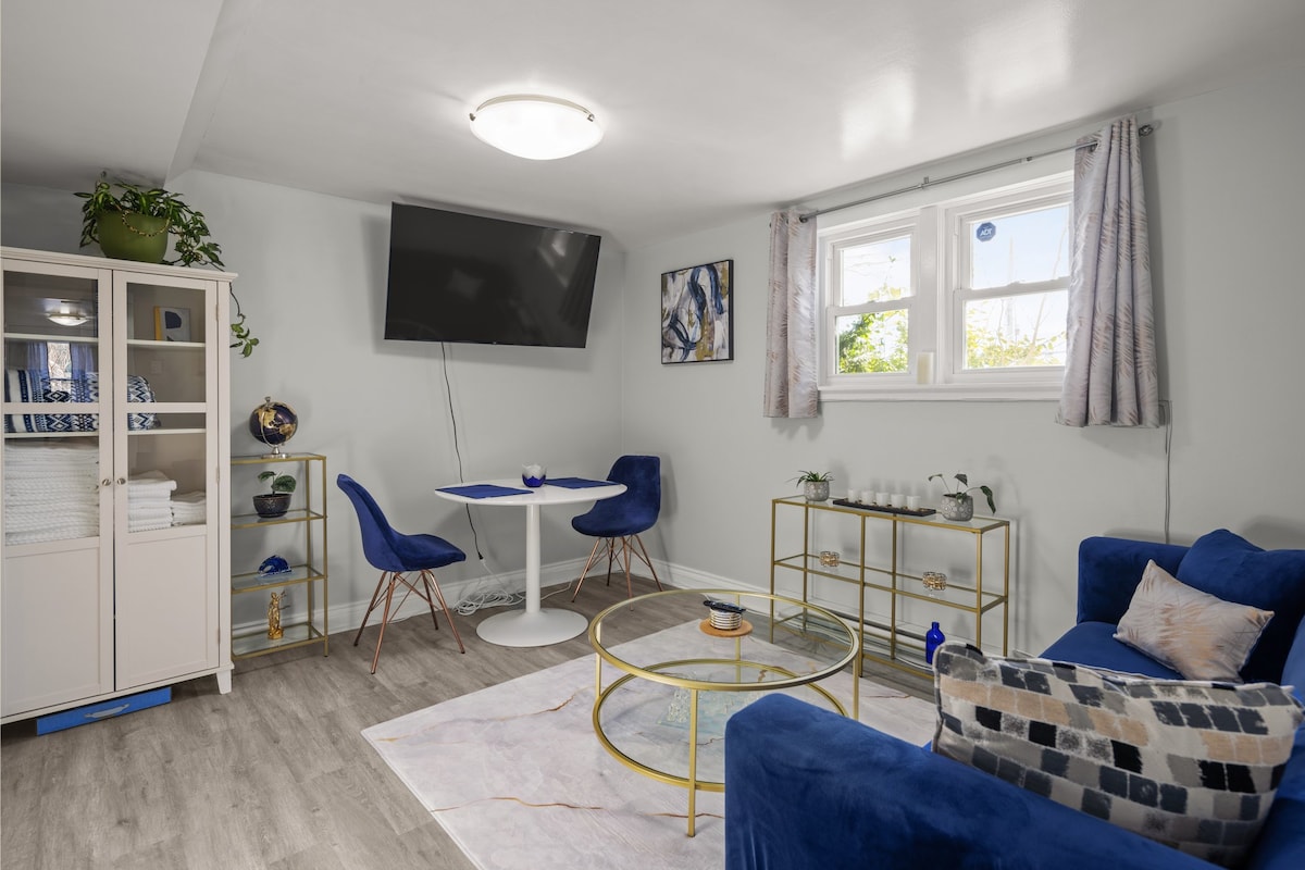A modern living space features a navy blue sofa, complemented by decorative cushions. A round glass coffee table sits on a patterned area rug. In the corner, a small dining table for two is set against a window, while a wall-mounted TV offers entertainment options.