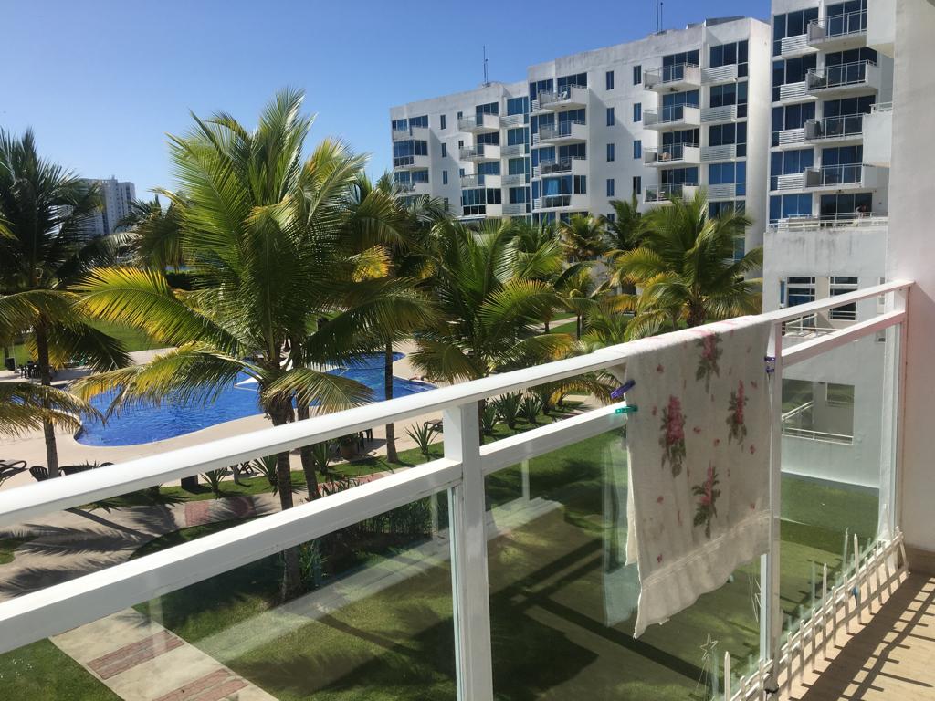 A balcony view is presented, featuring leafy palm trees and a pool area below. The light fabric hung on the railing adds a subtle touch to the scene, while modern apartment buildings are visible in the background under a clear blue sky.
