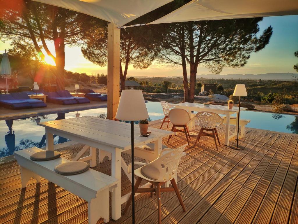 A spacious outdoor area features a large wooden dining table surrounded by white chairs, with a bench along one side. Sun loungers are positioned beside a pool, while tall trees and a setting sun create a serene backdrop.