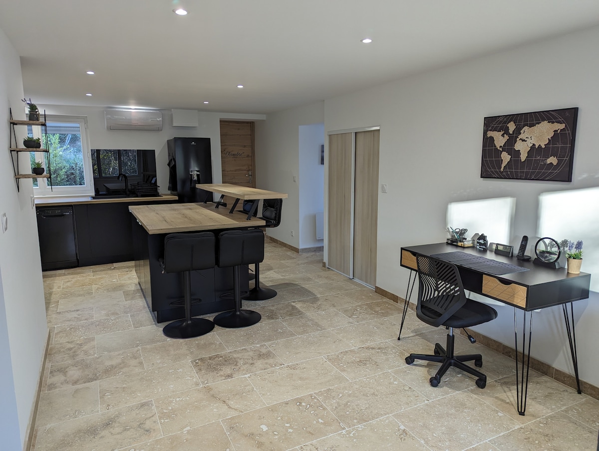 A modern and open kitchen area features a large island with bar seating. Dark cabinetry is complemented by light stone flooring. A workspace with a black desk and two chairs is positioned near a window, allowing natural light to enhance the space.