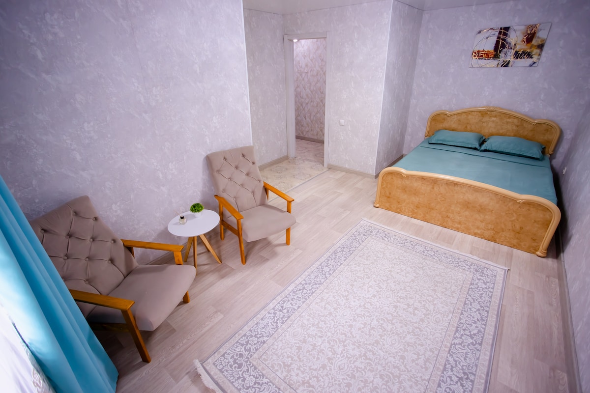 A softly lit bedroom features a spacious bed with light blue linens. Two beige chairs are positioned beside a small round table. A neutral-colored area rug anchors the space, while a doorway leads to another room, subtly framed by light blue curtains.